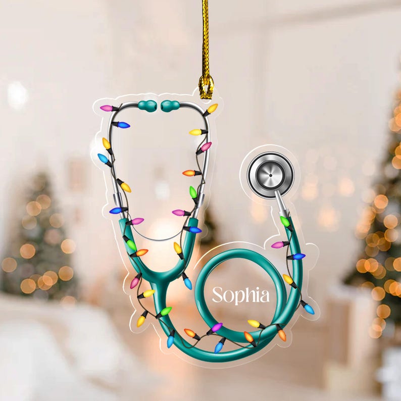May include: A teal stethoscope Christmas ornament with colorful lights. The ornament has the name "Sophia" on it and is hanging from a gold string. The background is blurred with Christmas trees.