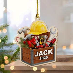 May include: Wooden Christmas ornament shaped like a toolbox, featuring tools, a yellow hard hat, and the name "JACK 2025". The ornament includes a red bow, candy canes, and greenery, with a gold hanging string.