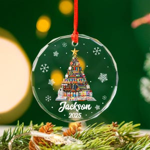 May include: A clear, round Christmas ornament with a red string. The ornament features a Christmas tree design made of art supplies, a gold star, snowflakes, and the name "Jackson" with the year "2025".