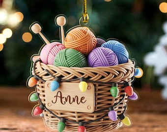 May include: A Christmas ornament shaped like a woven basket filled with colorful yarn balls and knitting needles. The basket is adorned with multi-colored Christmas lights and a wooden name tag that reads "Anne".