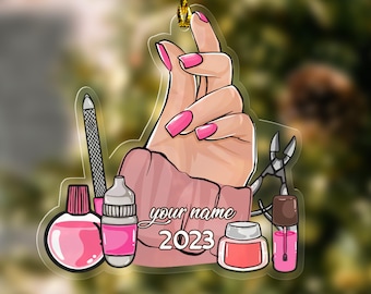 May include: A pink and white Christmas ornament featuring a hand with pink nail polish, nail clippers, and nail polish bottles. The ornament also includes the text "your name 2023".