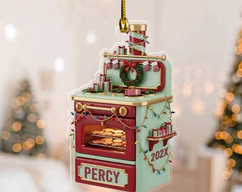 May include: A Christmas ornament shaped like a vintage oven, in red and mint green, with a gold trim. The oven door is open, revealing cookies. The ornament is decorated with a wreath, gifts, and string lights. The name "Percy" is on the front.