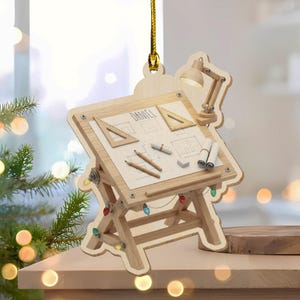 May include: Wooden Christmas ornament shaped like an architect's drafting table. The ornament features a miniature lamp, drafting tools, and a blueprint with the name "DANIEL". Colorful lights are strung along the base of the table.