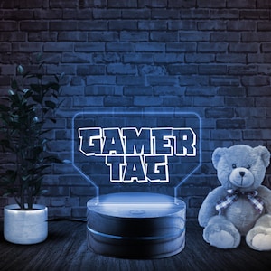 May include: A blue LED light with a wooden base displays the text "GAMER TAG" in a white font. The light is on a wooden surface next to a gray teddy bear.