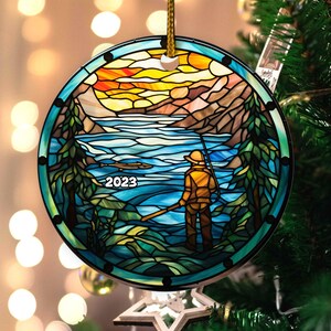 May include: A stained glass Christmas ornament featuring a fisherman standing in a lake with a mountain and sunset in the background. The ornament is blue, green, yellow, and orange. The year 2023 is written in the bottom left corner.