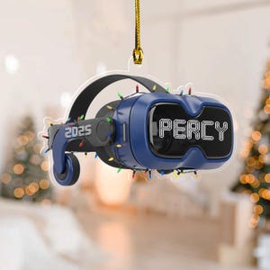 May include: A blue VR headset Christmas ornament with the name "PERCY" in white text. The ornament has the year "2025" printed on the side and is decorated with colorful string lights. A gold string is attached for hanging.
