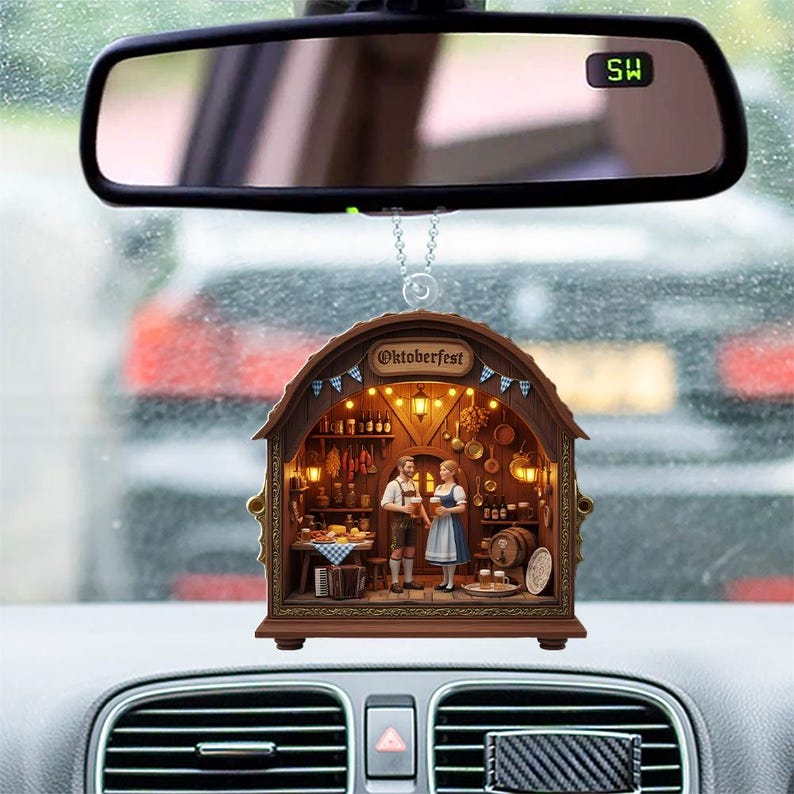 May include: A car rearview mirror ornament featuring a brown, arched wooden frame with the text Oktoberfest. Inside, a miniature tavern scene displays a man and woman in traditional attire, surrounded by shelves, barrels, and warm lighting.