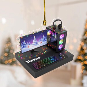 May include: A Christmas ornament featuring a gaming setup with a monitor displaying a winter scene, a computer tower, keyboard, mouse, and headphones. The name "Thomas" is on the mousepad. The setup is decorated with colorful lights.
