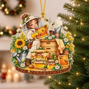 May include: A decorative ornament featuring a beekeeper in a white suit holding a honeycomb frame, surrounded by sunflowers, a beehive, a honey jar labeled Sophia, a honey dipper, and flying bees, all suspended by a gold cord for holiday display.