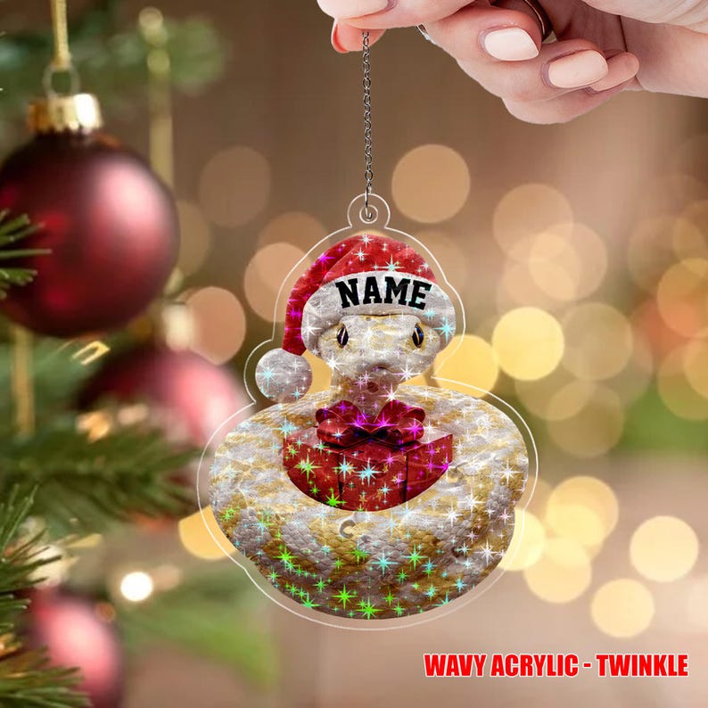 May include: A clear acrylic Christmas ornament shaped like a snake wearing a Santa hat with the word "NAME" on it. The snake has a red gift box on its belly. The ornament has a wavy texture and the text "WAVY ACRYLIC - TWINKLE".