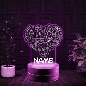 May include: A white heart-shaped night light with a science theme. The heart is filled with various science-related icons, including beakers, test tubes, molecules, and a microscope. The night light has a wooden base and a customizable name plate that reads "NAME".