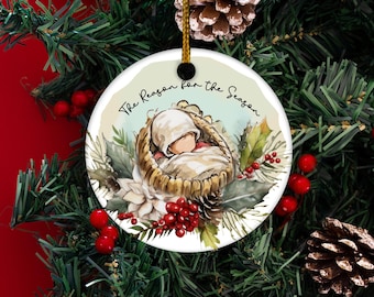 May include: A round Christmas ornament with the text "The Reason for the Season." The ornament features an illustration of a baby in a manger, surrounded by greenery and red berries. A gold string is attached for hanging.
