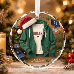 May include: A round, clear glass Christmas ornament featuring a green zip-up hoodie over a white t-shirt with the name BRIAN and a heart icon. It includes a Santa hat, computer icons, holly, snowflakes, and the year 2026 printed on it.