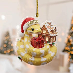 May include: A yellow snake Christmas ornament wearing a Santa hat with the name "Lucas". The snake is coiled around a red gift box and a gingerbread house. The ornament is hanging from a gold string.