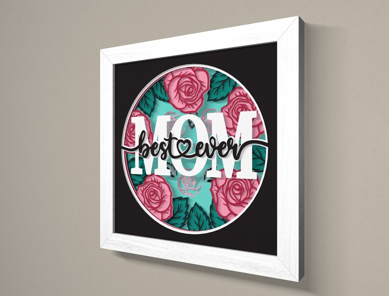 May include: White framed wall art with a black background and a circle in the centre. The circle has a turquoise background with pink roses and green leaves. The text "MOM best &hearts; ever" is in black lettering.