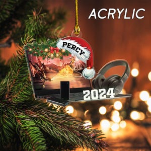 May include: Acrylic Christmas ornament featuring a laptop with a dragon scene, a Santa hat with the name "Percy", and black headphones. The year "2024" is displayed below. The ornament hangs from a gold string.