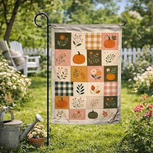 May include: A rectangular garden flag featuring a patchwork design of squares with illustrations of pumpkins, autumn leaves, wildflowers, and plaid patterns in shades of orange, green, cream, and brown, suspended from a black metal stand.