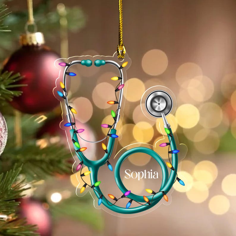 May include: A teal stethoscope Christmas ornament with the name "Sophia" printed on it. The ornament is decorated with a string of colorful Christmas lights. The ornament is hanging from a gold string in front of a blurred background of Christmas lights and a Christmas tree.