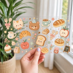 May include: A clear sticker sheet featuring various cartoon illustrations including a cat face, a bear with a party hat, a cup of coffee, a croissant, a slice of toast with butter, a cupcake, apples, bread, a jam jar, and a small cafe sign.