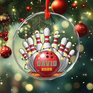 May include: A clear glass Christmas ornament featuring a bowling theme. The ornament has a red bowling ball with the name "DAVID" and the year "2025". White bowling pins with red stripes are arranged around the ball, with string lights.