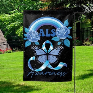 May include: A black garden flag featuring a blue butterfly, ribbon, and roses, with the word "ALS" above and "Awareness" below. A blue rainbow arches over the design. The flag is displayed outdoors on a metal stand.