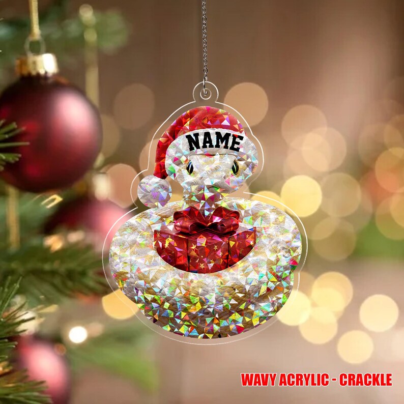 May include: A clear Christmas decoration featuring a Santa hat and a red gift box. The ornament has a wavy, crackle design and the word "NAME" is printed on the hat. It hangs from a silver chain.