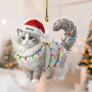May include: A cat ornament with a Santa hat and string lights. The cat is gray and white with blue eyes. The Santa hat has the word "Name" on it. The string lights are multi-colored. The ornament is hanging from a gold string.