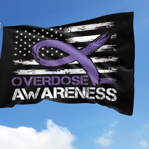 May include: A black flag with a distressed American flag design and a purple awareness ribbon. The words "OVERDOSE AWARENESS" are printed in white and purple. The flag is waving in the wind against a blue sky.