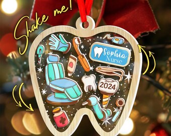 May include: Wooden tooth-shaped Christmas ornament with dental tools and a dental chair inside. The ornament has the text "Shake me!" and "Sophia Nurse 2024". It is hanging from a red ribbon, perfect for a dentist or dental hygienist.