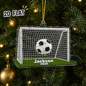 May include: A flat, clear acrylic Christmas ornament shaped like a soccer goal with a white net, a black and white soccer ball, and colorful string lights. The base features green grass with the text Jackson 2026 printed in white letters.