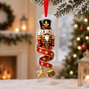 May include: A hanging nutcracker ornament featuring a figure in a red and green uniform with a black hat, attached to a red ribbon. A long red ribbon with gold stars spirals downward from the figure, ending with a gold star and a small drum.