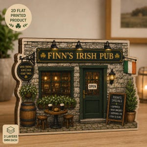 May include: A 2D flat printed wooden display of Finn's Irish Pub featuring a stone facade, a green door with an open sign, a window, a barrel, stools, and a table with two glasses. The sign reads Finn's Irish Pub, Good Friends Good Food Good Times.