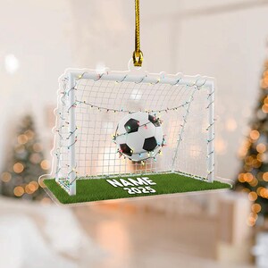 May include: A soccer-themed ornament featuring a soccer ball in a goal, decorated with string lights. The goal is set on a patch of green grass. The ornament has the text "NAME 2025".