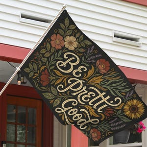 May include: Black flag with floral design and the words "Be Pretty Good" in cream script. The flag features flowers in shades of orange, yellow, and green. A sunflower is visible on the right side of the flag.