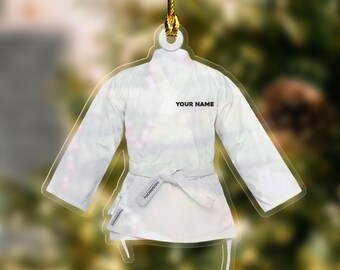 May include: A white karate uniform ornament with a gold string for hanging. The ornament features a karate gi with a belt and the text "YOUR NAME". The ornament is made of a clear material.