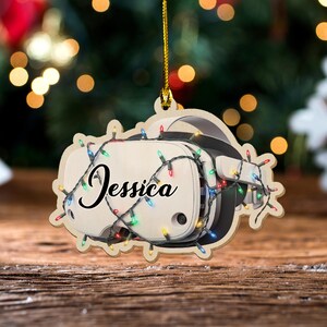 May include: Wooden Christmas ornament shaped like a VR headset, with the name "Jessica" in cursive. The ornament is decorated with a string of colorful Christmas lights. The background is blurred with Christmas lights.