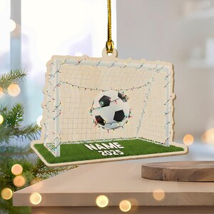 May include: Wooden soccer ornament featuring a soccer ball in a goal with a green field. The goal is decorated with string lights. The ornament has the text "NAME 2025" on the field. A gold string is attached for hanging.