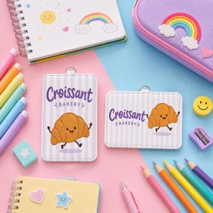 May include: Two rectangular badge holders with a light purple striped pattern, featuring a cartoon croissant character and the text Croissant Bakery. The items are surrounded by colorful stationery including markers, a notebook, and a pencil case.