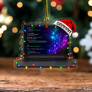 May include: A laptop-shaped ornament featuring a screen displaying computer code and a digital circuit pattern. A red and white Santa hat with the name GRAHAM printed on the brim sits on the corner. The ornament is outlined with colorful Christmas lights.