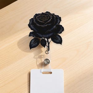 May include: A badge reel featuring a black rose with gold-toned outlines and subtle gold speckles on the petals. The rose is attached to a clear plastic strap and a circular metal housing, which connects to a plain white rectangular ID card holder.