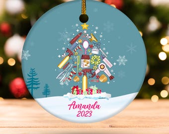 May include: Round Christmas ornament with a blue background and a Christmas tree design made of salon tools. The ornament has the name "Amanda" and the year "2023" written in pink script. Snowflakes and a small Christmas tree are also visible.