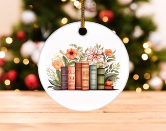 May include: A white, round Christmas ornament featuring a watercolor illustration of a row of books with floral accents. The books are in various colors, and the flowers are peach, pink, and white.