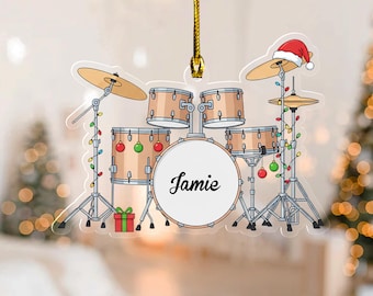 May include: A clear acrylic Christmas ornament shaped like a drum set. The drum set has a Santa hat on one cymbal and is decorated with Christmas lights and ornaments. The name "Jamie" is written on the bass drum.