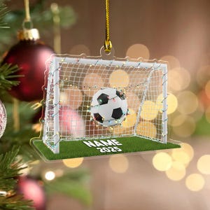May include: A clear acrylic soccer-themed Christmas ornament. The ornament features a soccer ball in a net, with a green base and the text "NAME 2025". The net is decorated with colorful lights, and is suspended by a gold string.