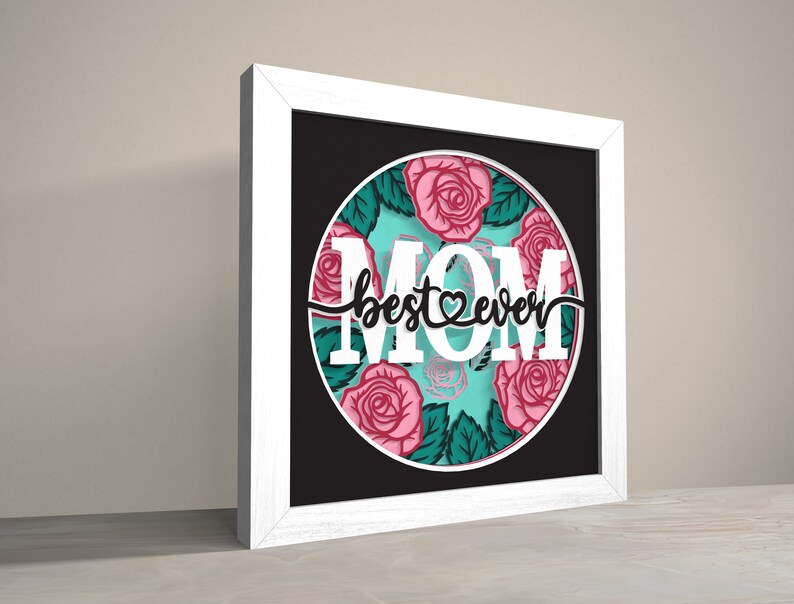 May include: White framed picture with a black background and a circle in the centre. The circle has a turquoise background with pink roses and green leaves. The text "best ever" is written in black with a heart in the middle of the word "best". The word "MOM" is written in white letters around the circle.