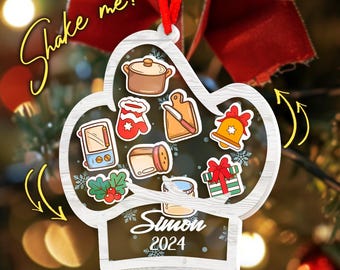 May include: A personalized Christmas ornament with a kitchen theme. The ornament is shaped like a chef's hat and features various kitchen-related illustrations, the name "Simon" and the year "2024". The ornament is hanging from a red ribbon.