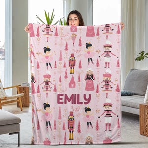 May include: Pink fleece blanket with a repeating pattern of a ballerina, a nutcracker, and a Christmas tree. The blanket has the name "EMILY" printed in a large font.