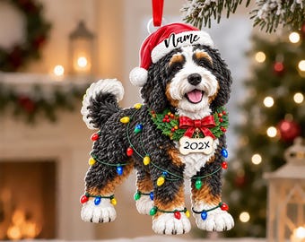 May include: A Bernese Mountain Dog Christmas ornament featuring a dog wearing a red Santa hat with the text Name, a green wreath with a red bow, and a bone-shaped tag labeled 202X. The dog is wrapped in colorful string lights and hangs by a red ribbon.