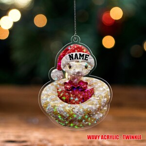 May include: A clear acrylic Christmas ornament shaped like a snake wearing a Santa hat. The ornament has the word "NAME" on the hat and a red gift box. The text "WAVY ACRYLIC - TWINKLE" is at the bottom.
