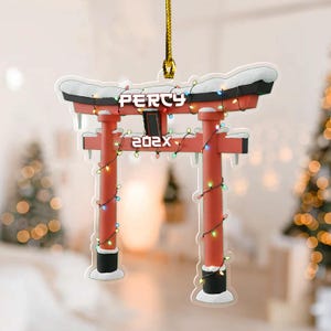 May include: A red Torii gate Christmas ornament with the text "PERCY 202X". The ornament is decorated with a string of colorful lights and snow. It hangs from a gold string.
